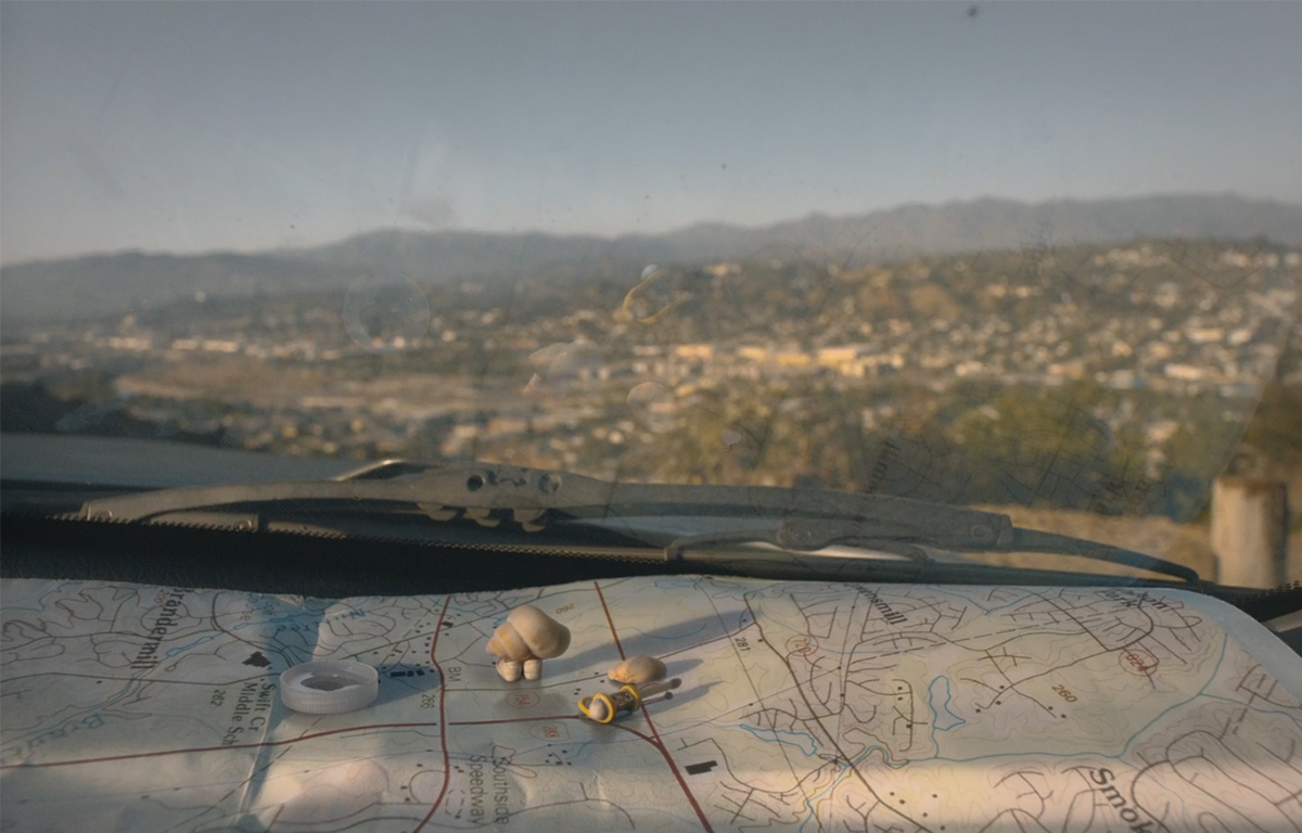 Marcel the Shell standing on top of a map on the dashboard of a car, looking at a view of a city from a high view point.