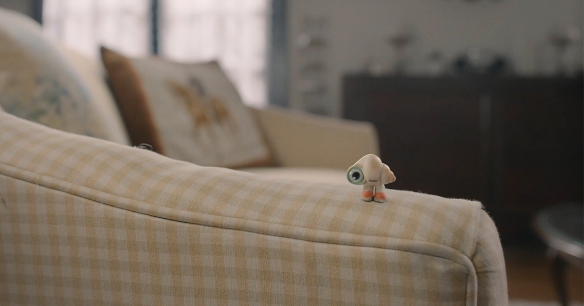 Marcel the Shell, a tiny shell with one eye and pink shoes on, standing on top of the arm of a couch in a living room.